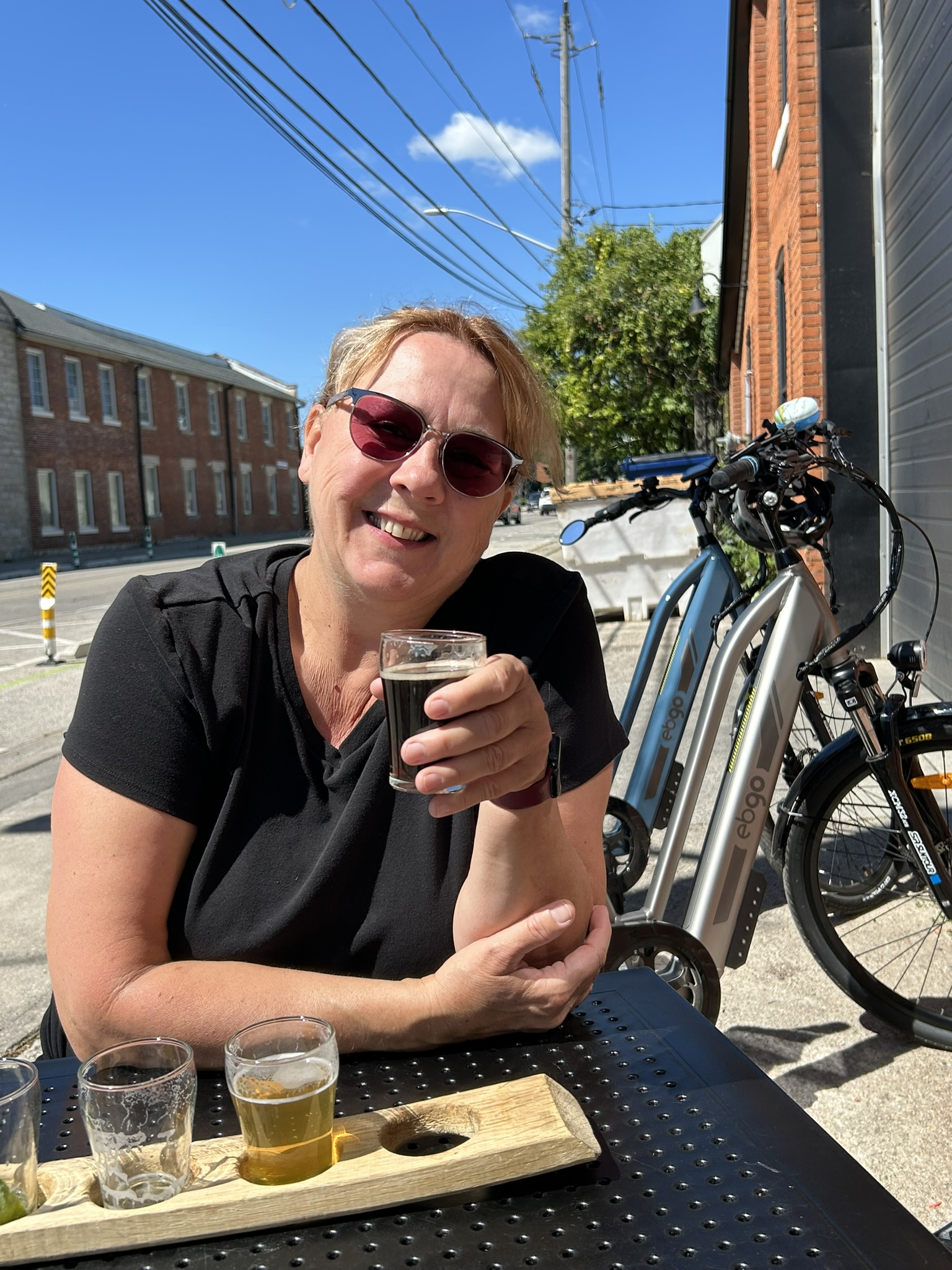 Sonya smiling on a patio with a beer flight, e-bike parked behind her on a sunny day